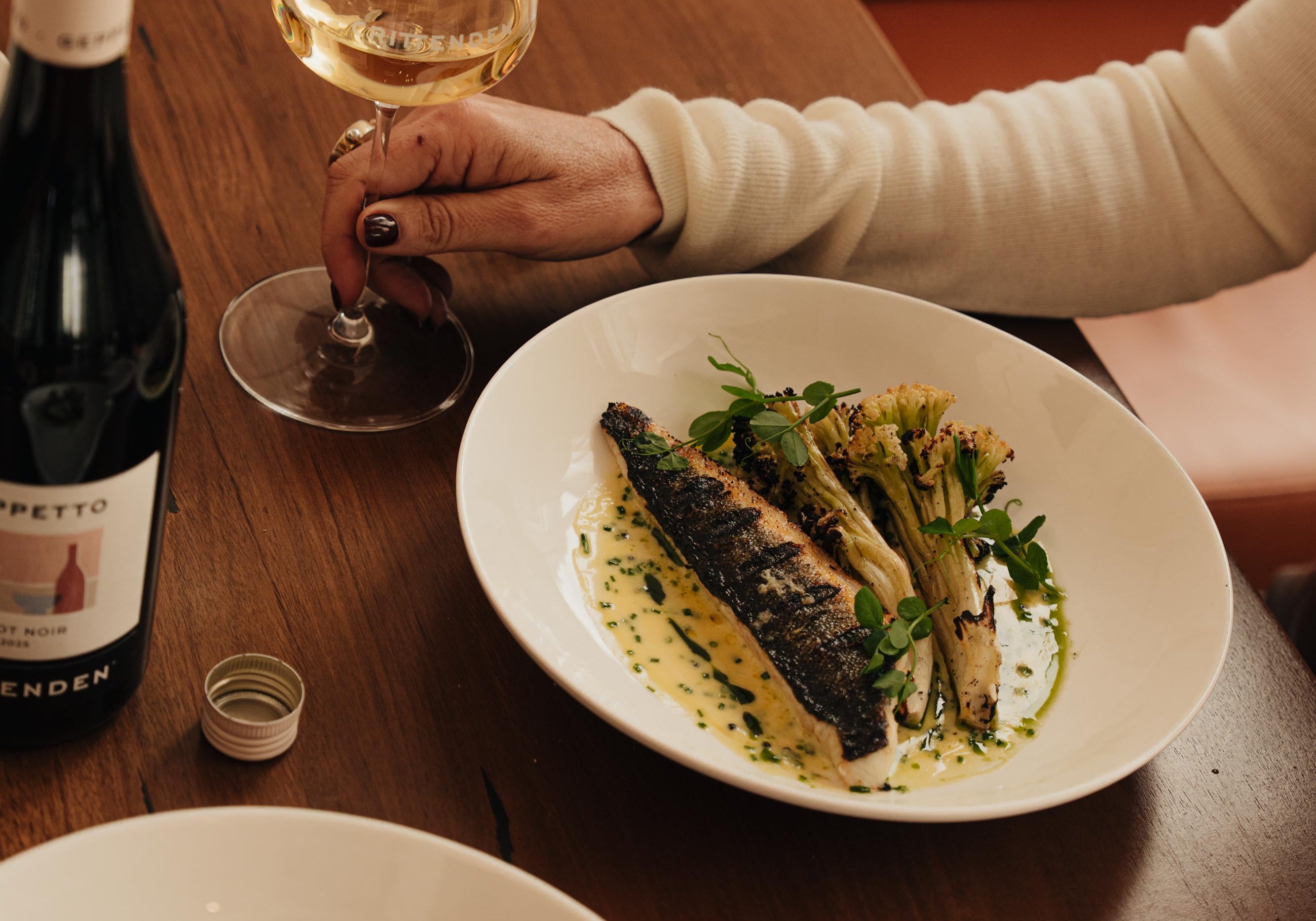A fish main with greens and a glass of white wine on a restaurant table, beside a bottle of Geppetto