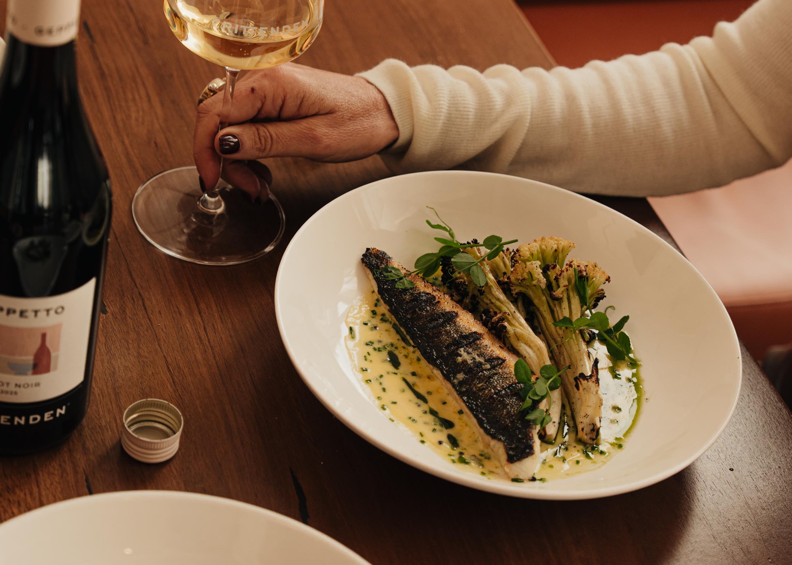 A fish main with greens and a glass of white wine on a restaurant table, beside a bottle of Geppetto