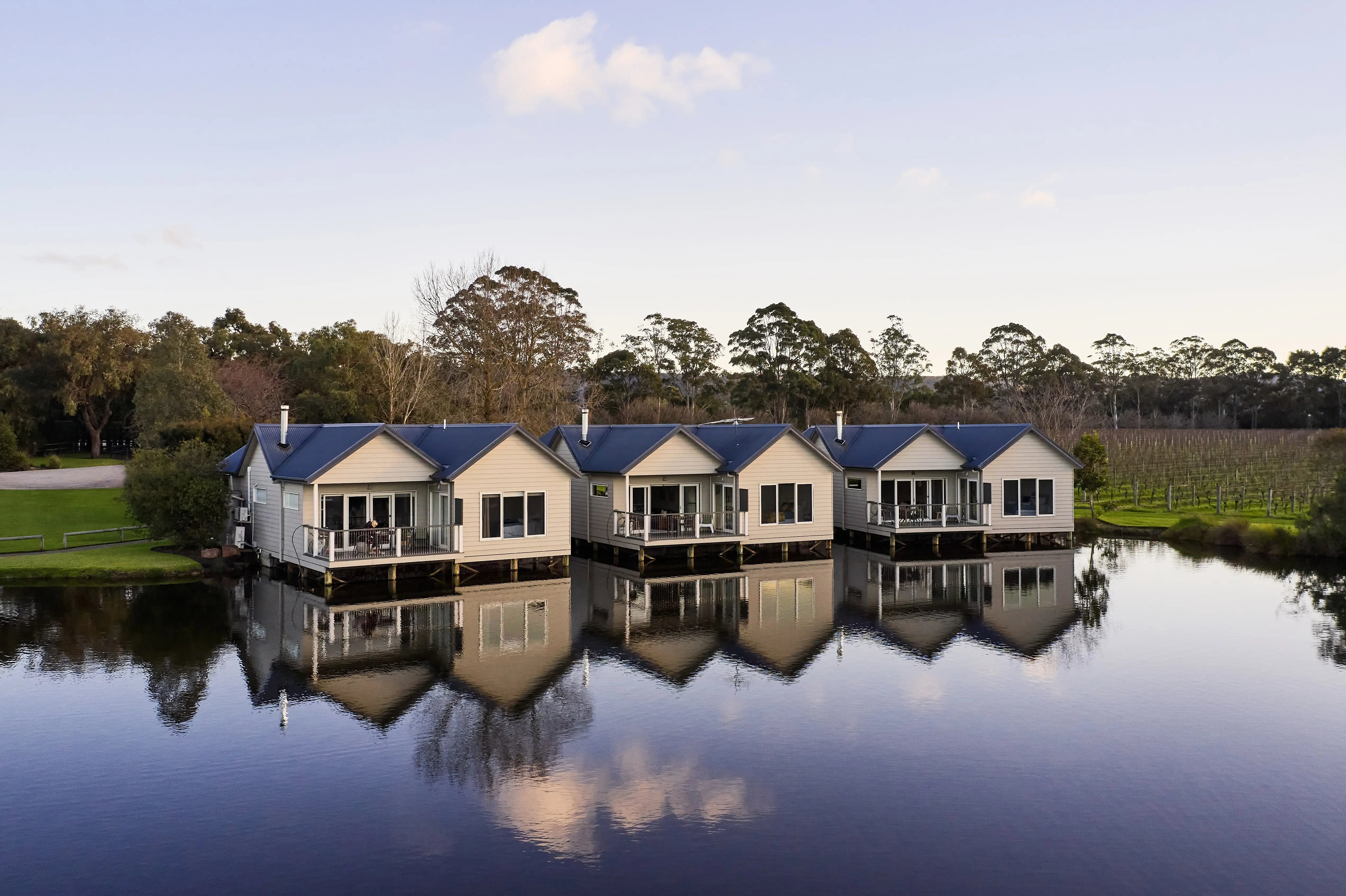 Row of Lakeside Villas reflected in a calm body of water with trees and vineyard in the background.