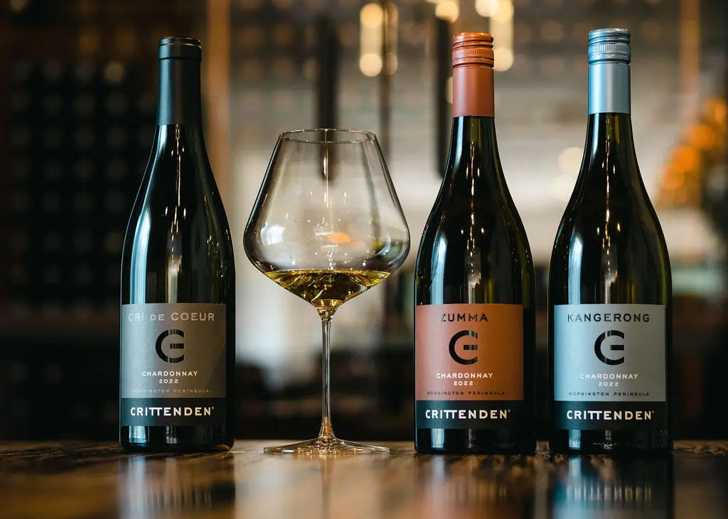 Three different Crittenden Chardonnay wine bottles and a glass of white wine on a wooden table.