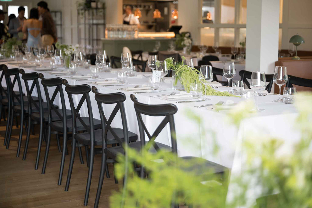 A long table set with white linen, glassware and black chairs for a reception