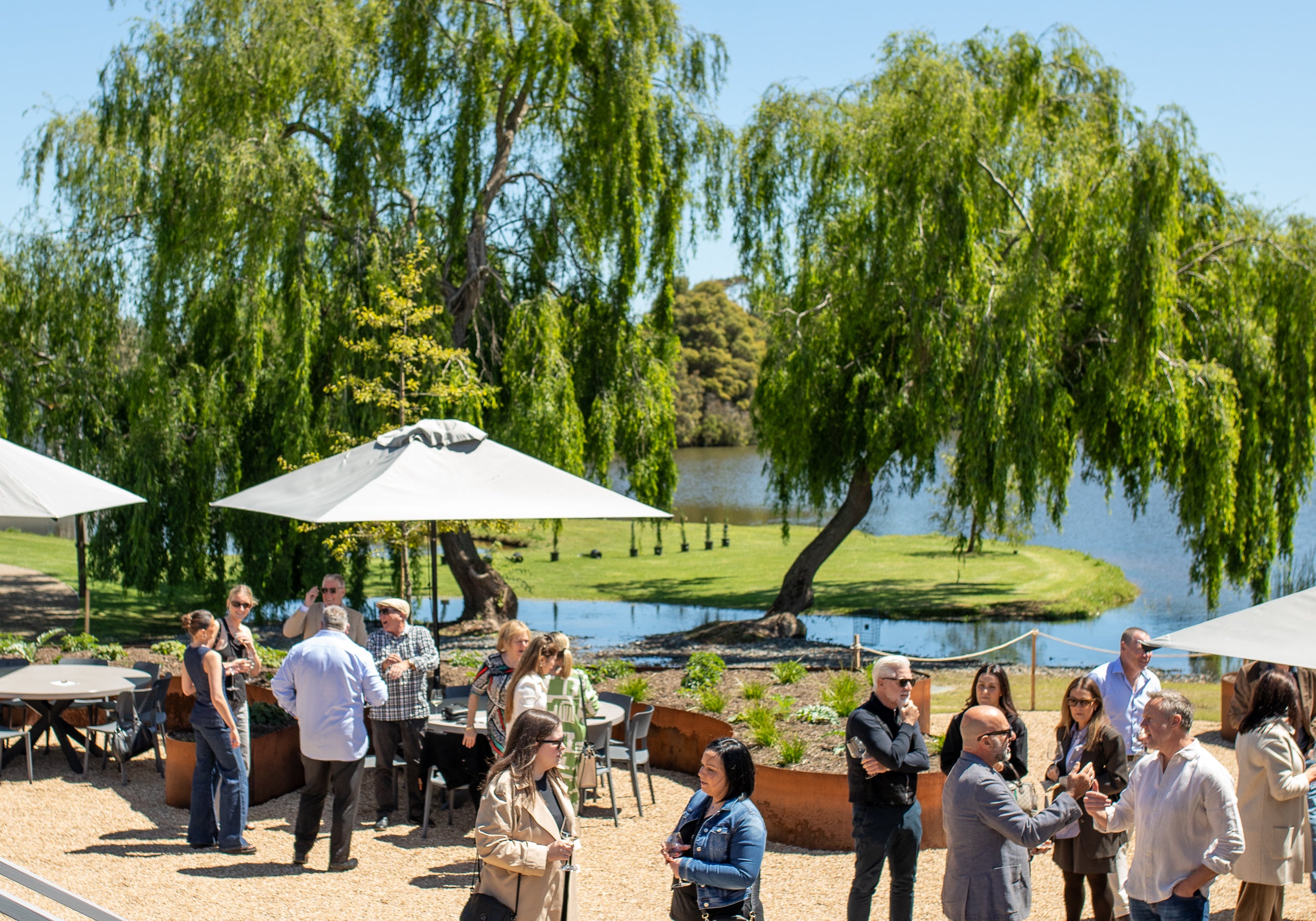 Guests gathered on the gravel by the lake under willows and umbrellas
