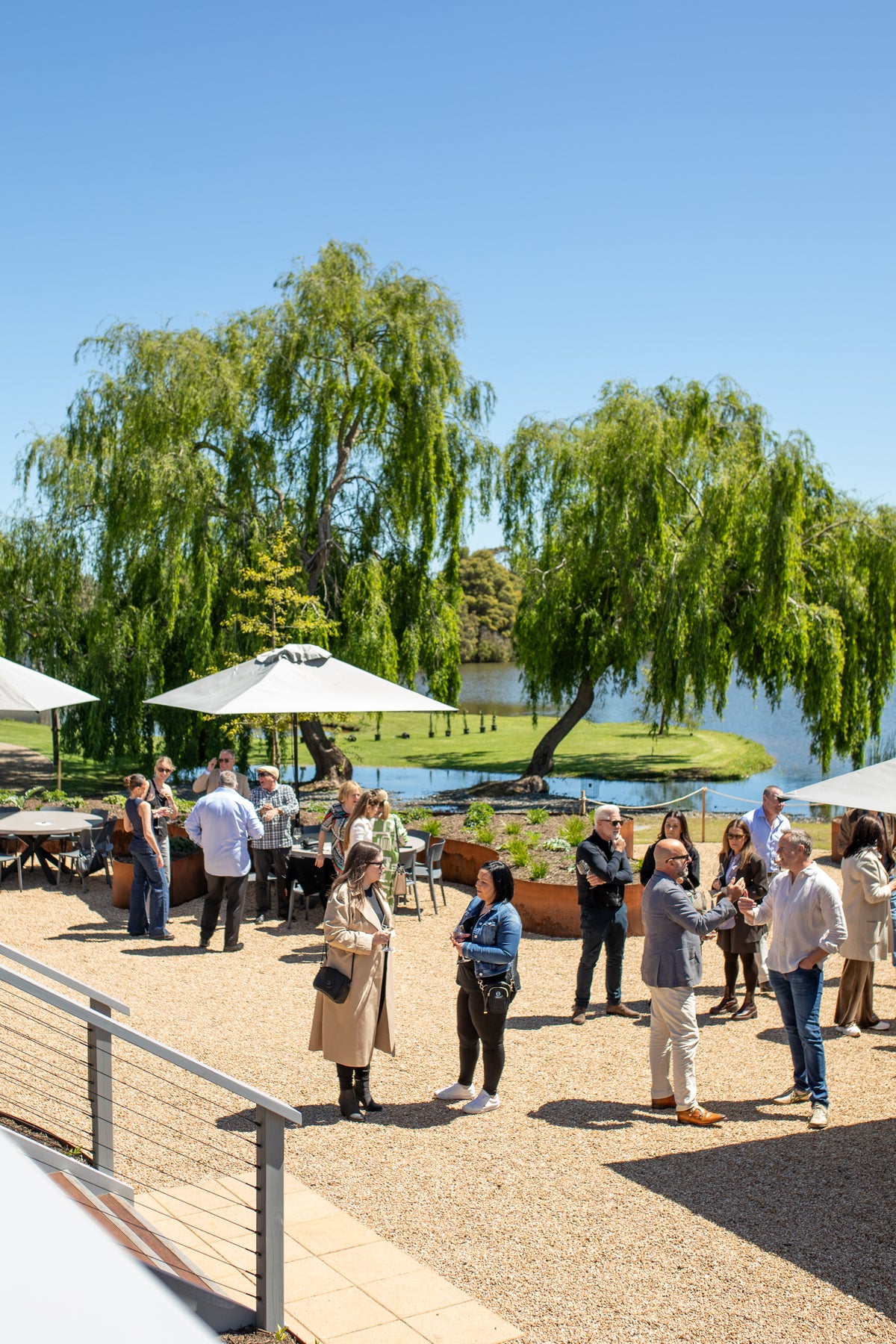 Guests gathered on the gravel by the lake under willows and umbrellas