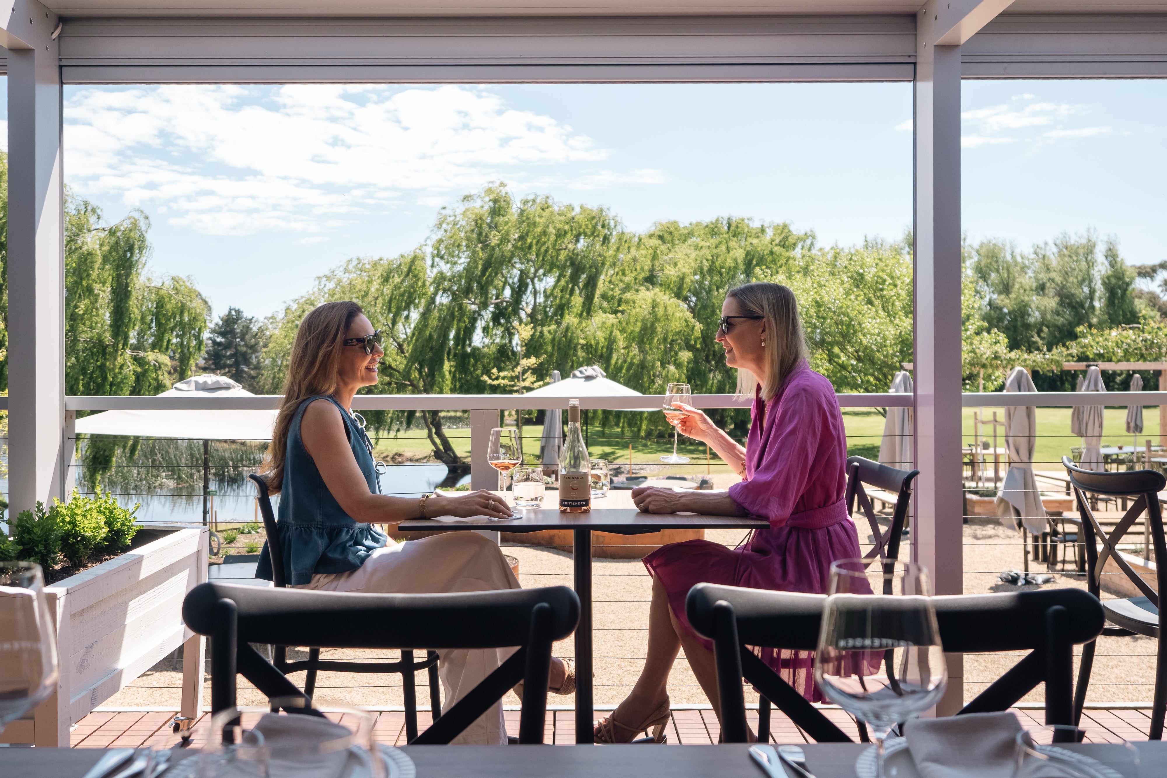 Two women sharing a bottle of wine at a table on a covered deck overlooking a lake and willow trees