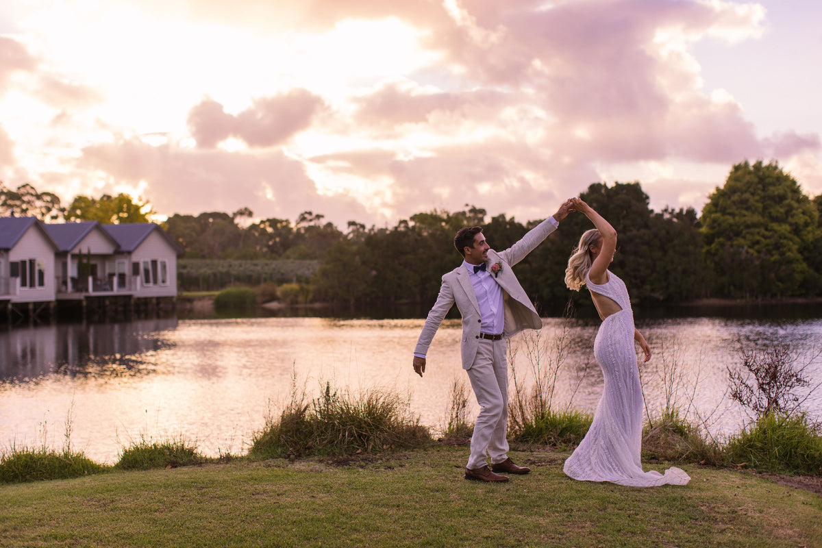 A bride and groom dancing on the lawn beside a lake at sunset, with villas over the water behind