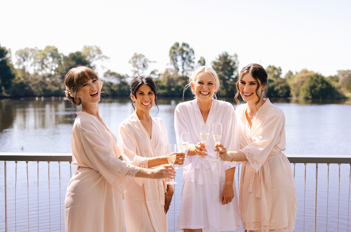 A bride and three bridesmaids in robes raise glasses of sparkling wine on a deck over the lake
