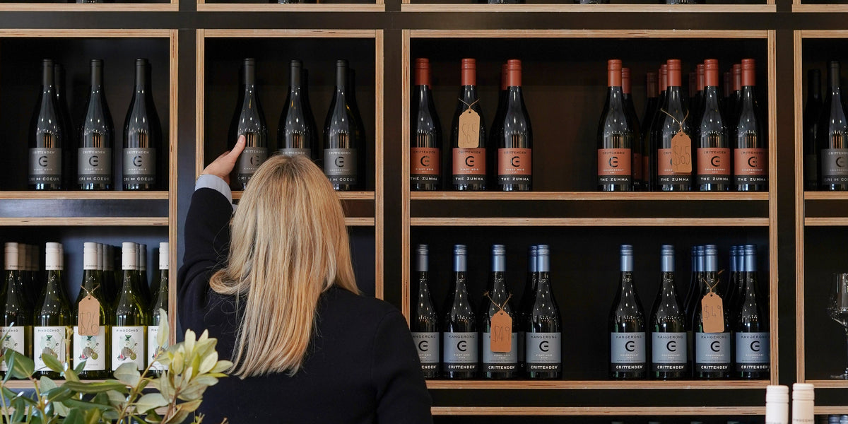 A woman with blonde hair, seen from behind, reaches toward a bottle on a wooden wine display shelf filled with other wine bottles.