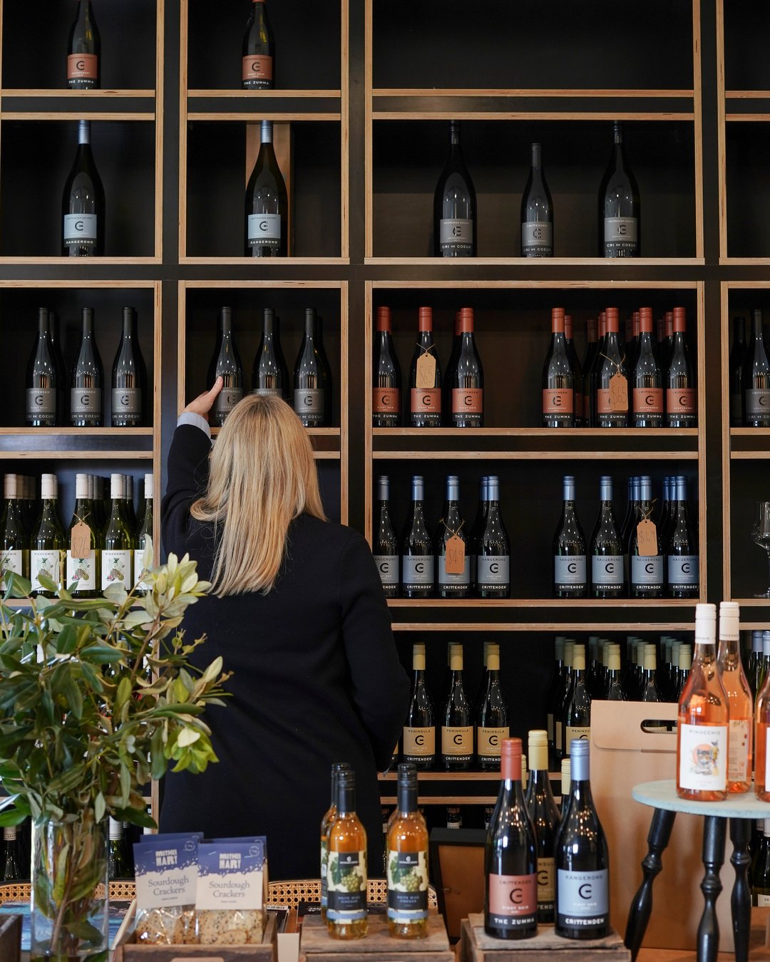 A woman reaches for a bottle on the cellar door's wall of wine shelves, with local produce on the table below