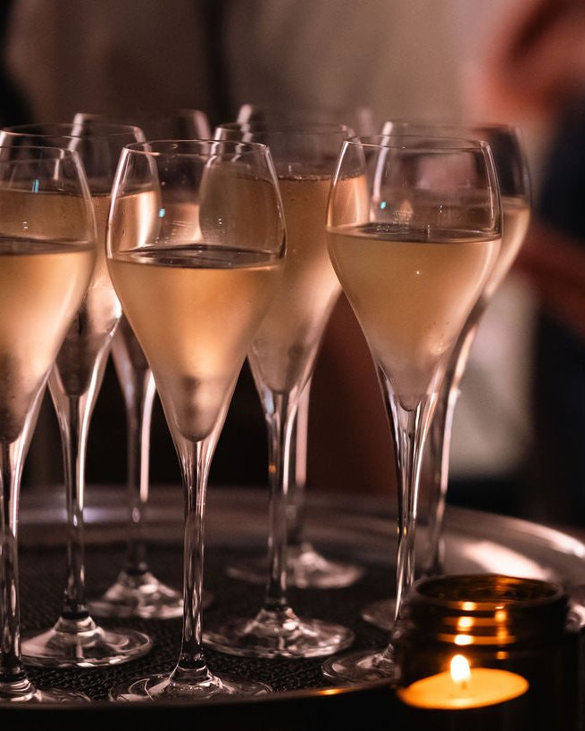 A close-up, low-angle shot of several champagne flutes filled with champagne on a tray, with a lit candle in the foreground and a softly blurred person in the background.