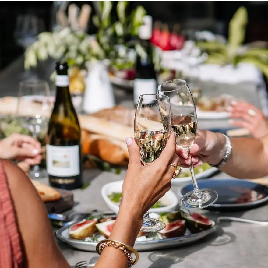 Two people clinking champagne glasses at a festive outdoor table setting with food and wine bottles.
