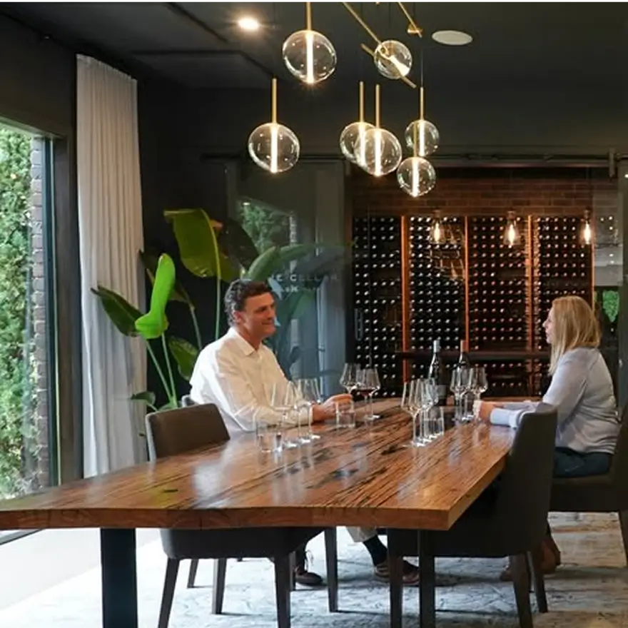 Two people sitting at a wooden table with wine glasses in a dimly lit room with wine bottles in the background.