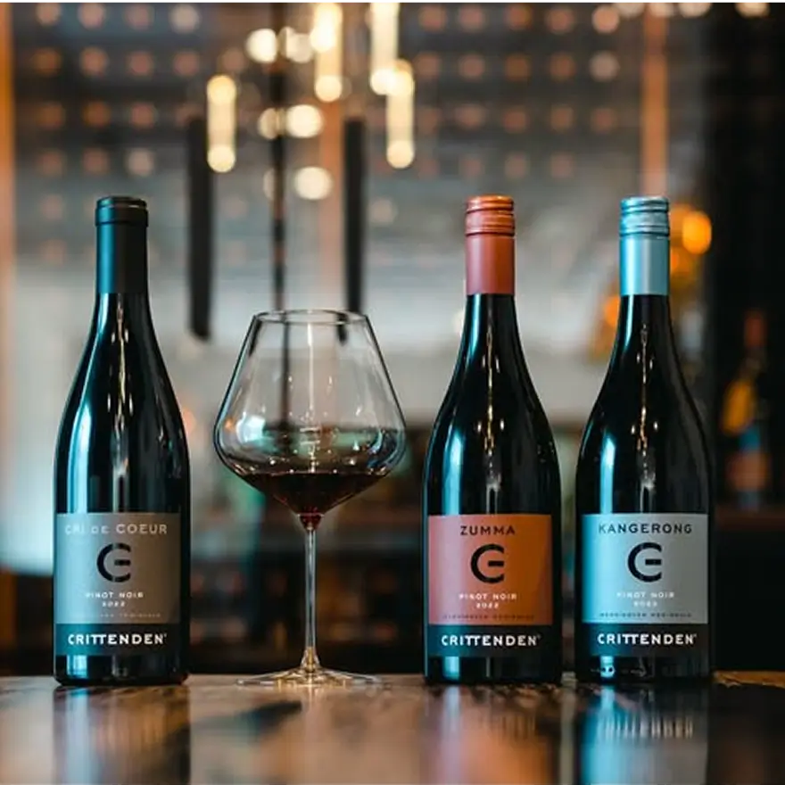 Three wine bottles with labels on a bar counter, one with a glass of red wine.
