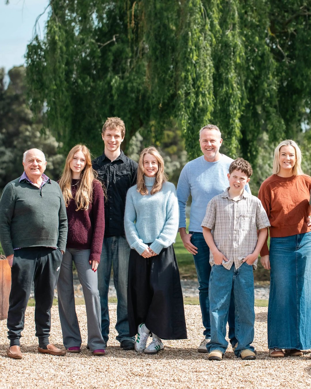 A group of 8 Crittenden family members together outdoors in front of a large weeping willow tree. The group is dressed in casual, comfortable clothing suitable for a family portrait, with gravel on the ground and garden elements in the background.