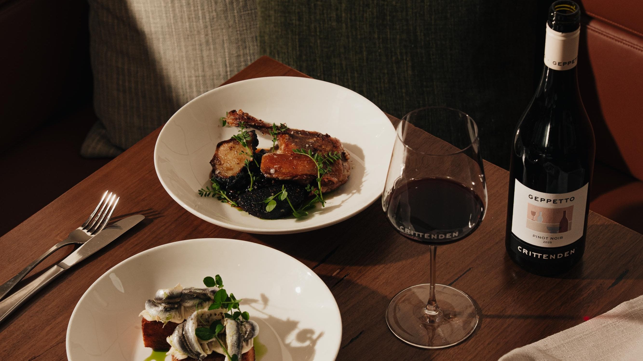 A dining table setting featuring a bottle of Cristero Geppetto red wine, a filled wine glass, and two plated dishes of food, captured in warm, moody ambient lighting.