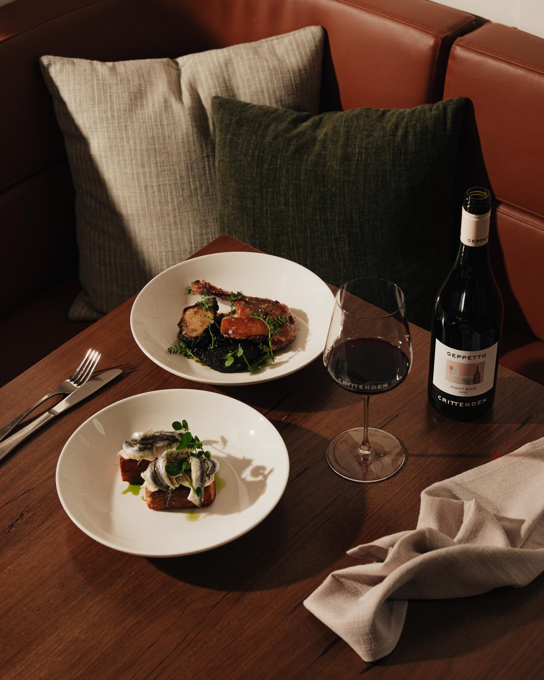A styled dining table scene with two white plates of food, a filled wine glass, a dark wine bottle, and a beige cloth napkin on a wooden table, set against a brown leather booth with pillows.