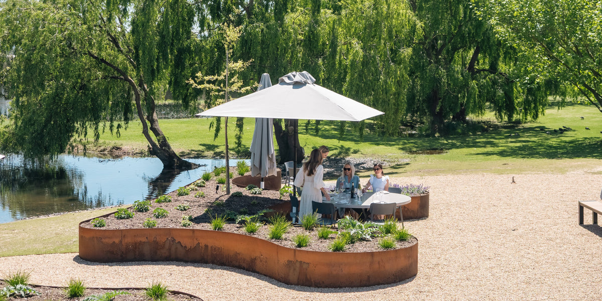 Crittenden Terrace area beside a pond with a large white umbrella, rust-colored metal planter beds, and several people sitting and standing in a landscaped park setting.