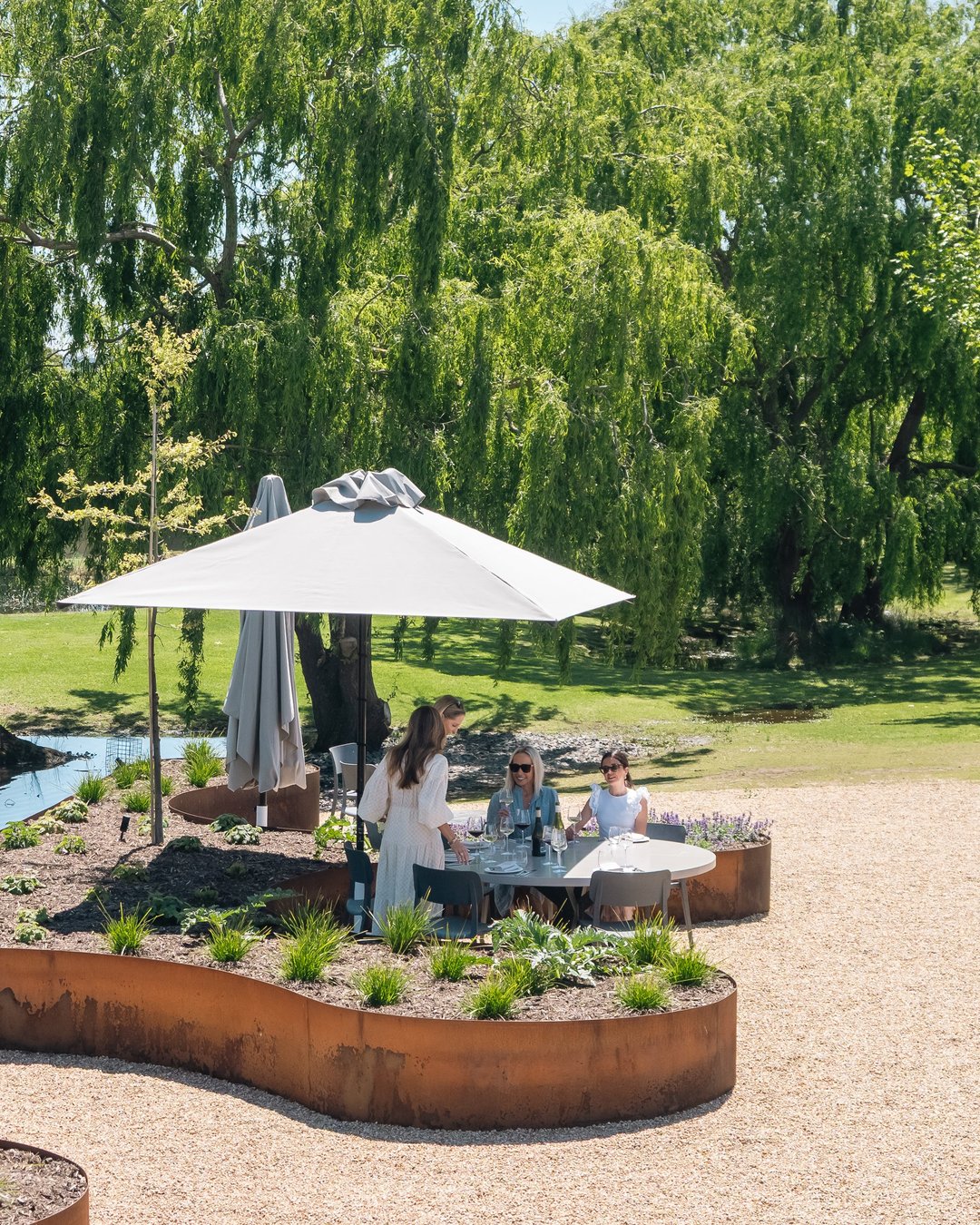 Crittenden Terrace seating area with a large white patio umbrella, round metal tables, and three people sitting and standing in a landscaped garden with gravel ground, rust-toned planter beds, and a weeping willow tree in the background.