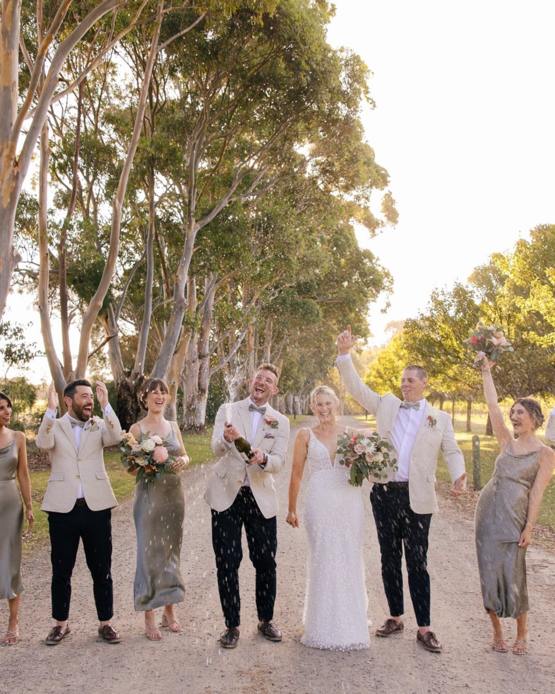 A bride, groom and wedding party cheer as champagne sprays on a path lined with gum trees
