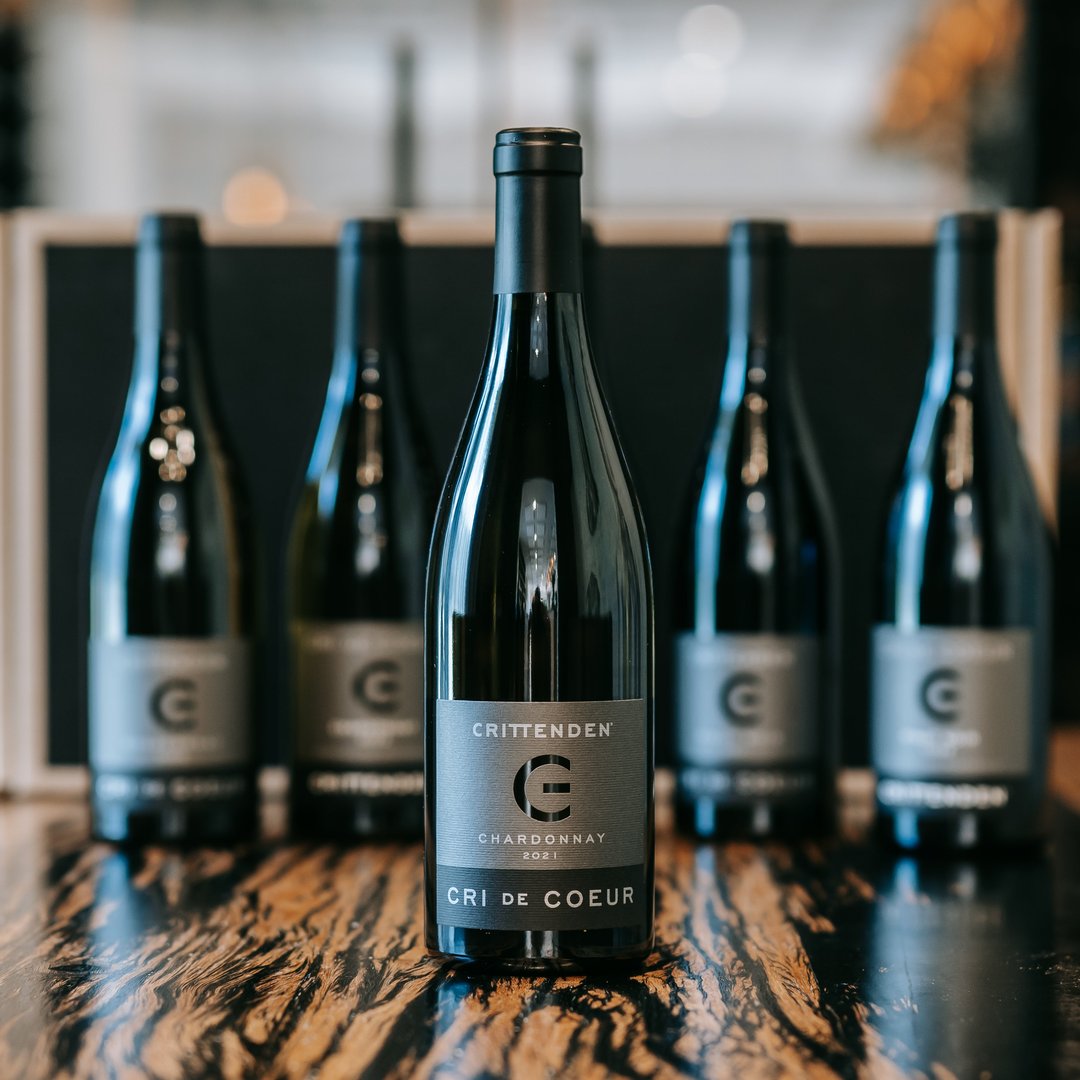 A bottle of Crittenden Chardonnay 2021 in the foreground, with four matching bottles lined up behind it on a dark wooden surface.