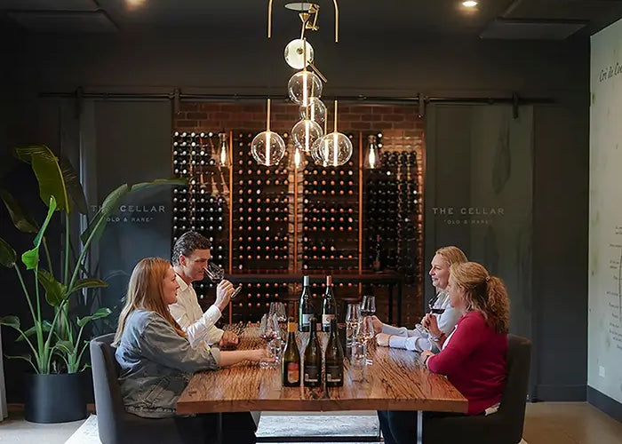 Four guests tasting wine around a timber table in the private tasting room, with a wall of bottles behind them